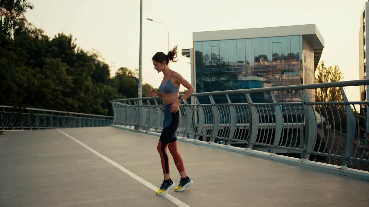 A sports girl in a sports summer uniform runs in place and warms up before a morning run against the background of a bridge in the city