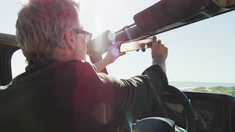 Hombre-Caucásico-Con-Gafas-De-Sol-Sentado-En-El-Coche-Ajustando-El-Espejo-Retrovisor-En-Un-Día-Soleado-En-La-Playa