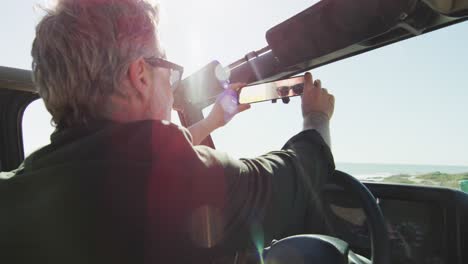 hombre caucásico con gafas de sol sentado en el coche ajustando el espejo retrovisor en un día soleado en la playa