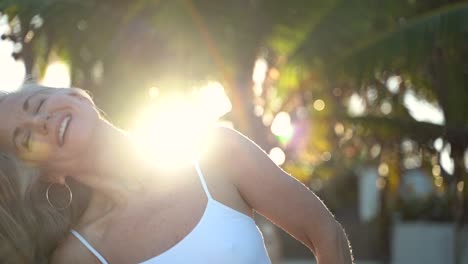 Sexy-Zeitlupe-Einer-Reifen-Blonden-Frau-Mit-Sonne,-Die-Durch-Palmen-Scheint,-In-Die-Kamera-Lächelt-Und-Mit-Ihren-Augen-Flirtet