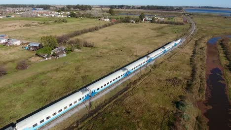 Tren-Deslizándose-Por-Vías-Cerca-De-Un-Río-Sinuoso-Con-Casas-Rurales-Al-Fondo,-En-Las-Afueras-De-Buenos-Aires.