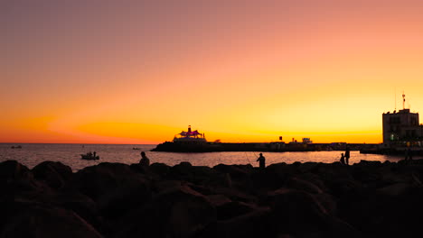 Pescadores-De-Silueta-En-El-Muelle-Y-La-Costa-Durante-La-Puesta-De-Sol-Naranja-Lanzando-Sedal-En-Mar-Abierto-En-El-Fondo-Es-La-Ciudad-Y-El-Puerto-De-La-Isla-Gran-Canaria-Puesta-Del-Sol-4k-Captura-En-Cámara-Lenta-A-60fps