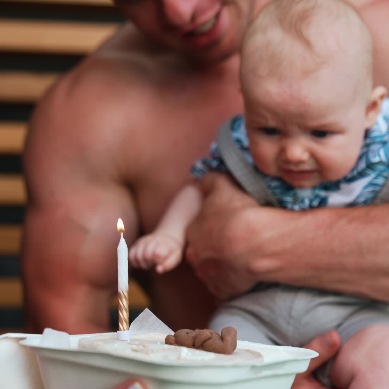 Küçük yıldönümünü kutlayan genç aile. Tanınmayan anne pastanın üzerinde bir mum yakarken, baba bir bebek tutuyor.