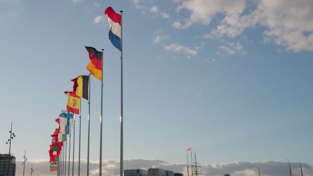 Colorful national flags line the harbor, led by Dutch, German and Spanish flags under a partly cloudy sky. Shot in Amsterdam, Netherlands (Nederland)
