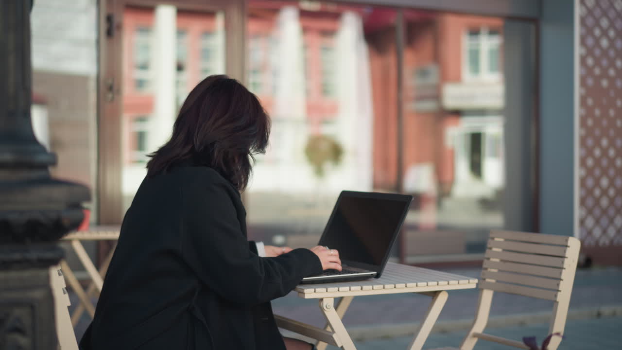 Lady working on laptop outdoors with hair covering her face, coffee cup on table beside her, decorative black pillar nearby, and blurred background featuring urban building and glass