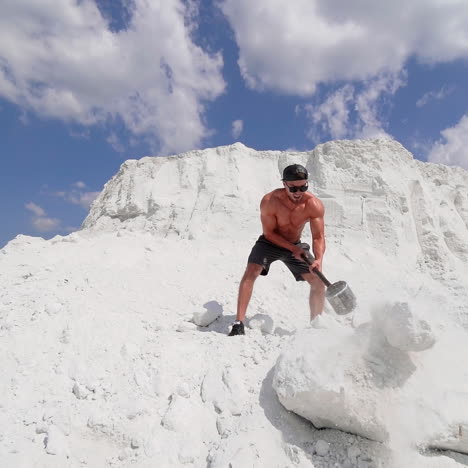 muscle athlete strongman in a cap and sunglasses man hits a huge white rock with a hammer in the white sandy mountains or in a quarry. slow motion. sunny day . white landscape.