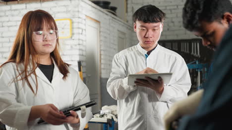 실험실 코트를 입은 학생들은 엔진에서 도구 사용을 보여주는 메카닉을 관찰하면서 태블릿에 메모를 취합니다.