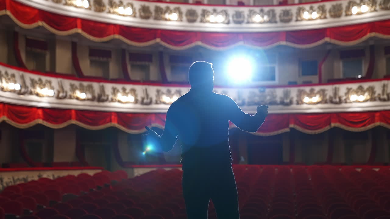 Announcer on a stage in theater. Bright round light on one person. Man in black suit performs to the auditorium. View from the back.
