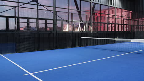 Empty indoor padel tennis court with blue flooring and large windows, copy space
