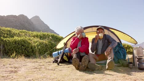 Happy-senior-biracial-couple-sitting-at-tent-in-mountains-and-drinking-coffee,-in-slow-motion