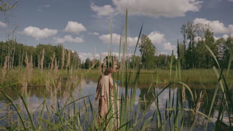 a woman is walking by the pond as the camera is following her with tall grasses in the foreground, colour graded, slow motion