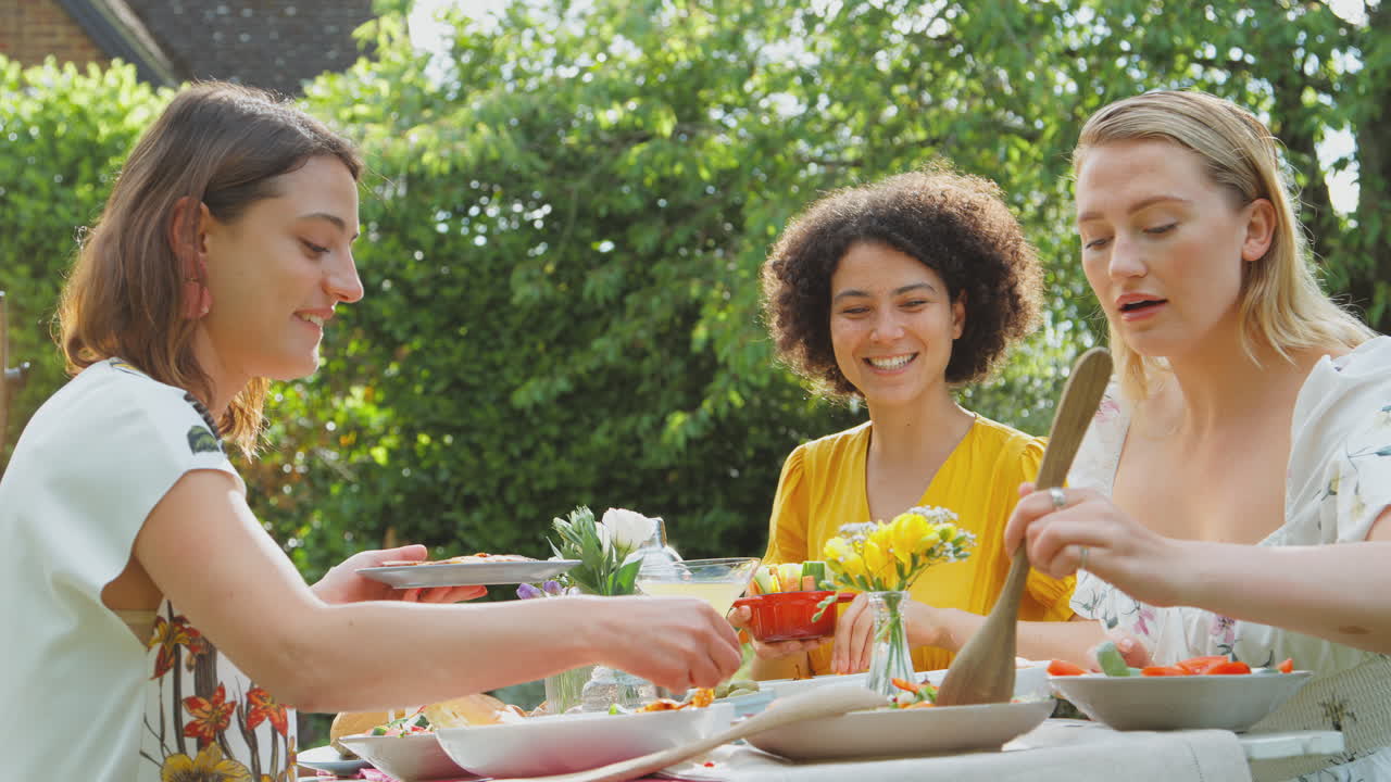 tres amigas sentadas al aire libre en el jardín de verano en casa bebiendo cócteles y comiendo comida