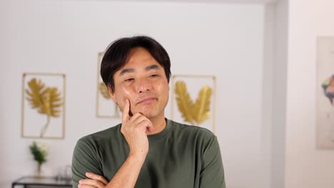 Middle-aged Asian man stands indoors, touching his face in a thoughtful pose. Soft, even lighting and minimal camera movement create a calm, introspective mood