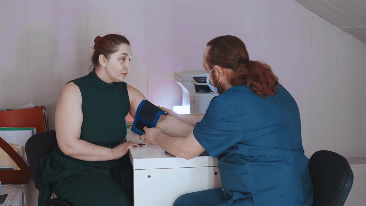 Paziente donna bianca con controllo della pressione sanguigna a domicilio da parte di un paramedico uomo bianco, bracciale blu sul braccio, accogliente ufficio domestico, stampante e documenti sulla scrivania, interazione calma e di supporto e rassicurazione attenta
