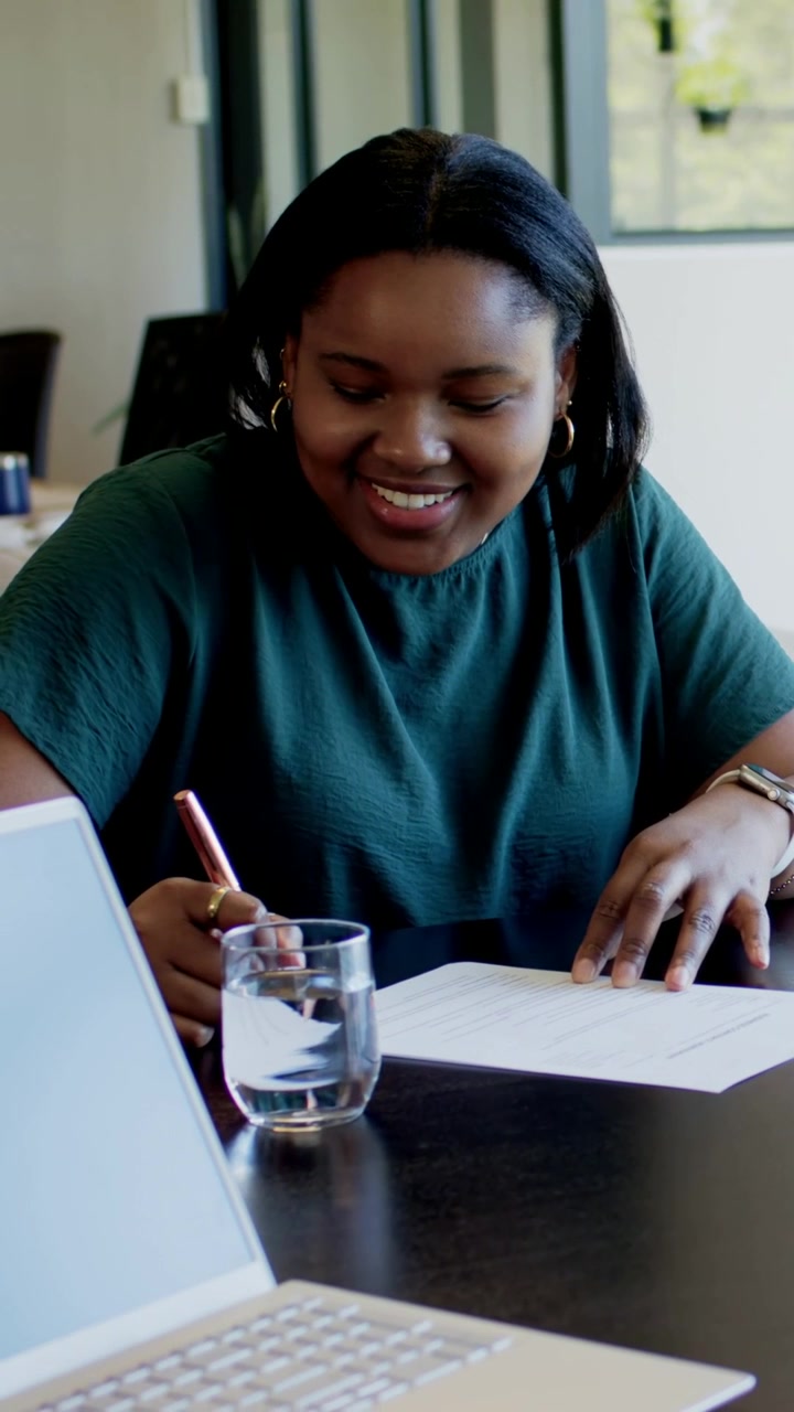 Vertical video: Beginning form woman in green top writing paper with pen at table, laptop nearby