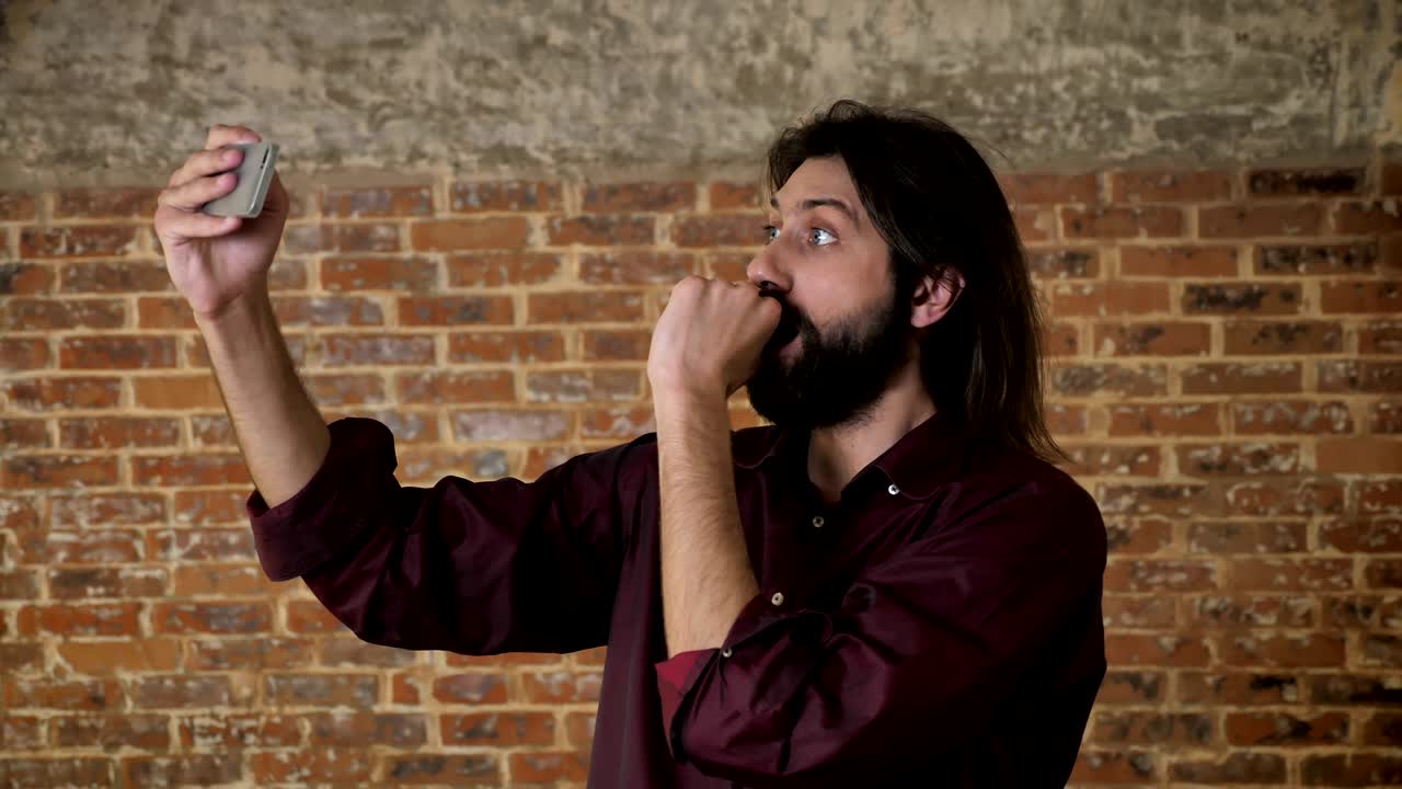 joven atractivo hombre moreno con barba está haciendo video en el teléfono inteligente, tonto, fondo de ladrillo, concepto de comunicación