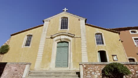 Small-church-in-the-sun-of-France-with-colored-facade-and-cross-on-the-top-closed-door