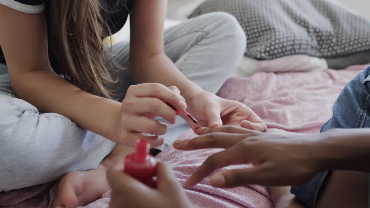 Diverse teenage female friends sitting on bed and painting each other's fingernails
