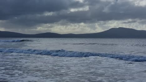 following rough stormy shimmering waves breaking under misty snowdonia mountain range with sunrays shining through overcast clouds