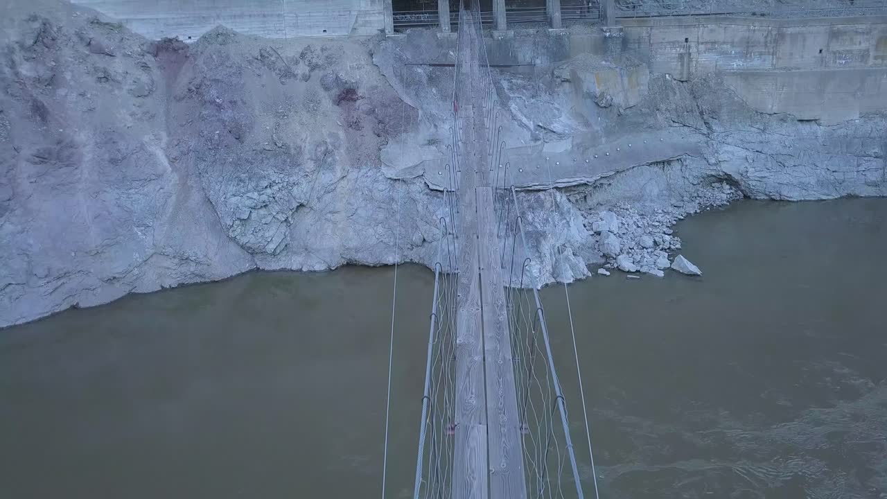 Muddy canyon river swirls below narrow suspension foot bridge in shade