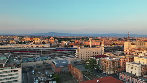 Forwards-fly-above-town-development-near-train-station-in-golden-hour.-Modern-passenger-trains-moving-on-railway-tracks.-Rome,-Italy