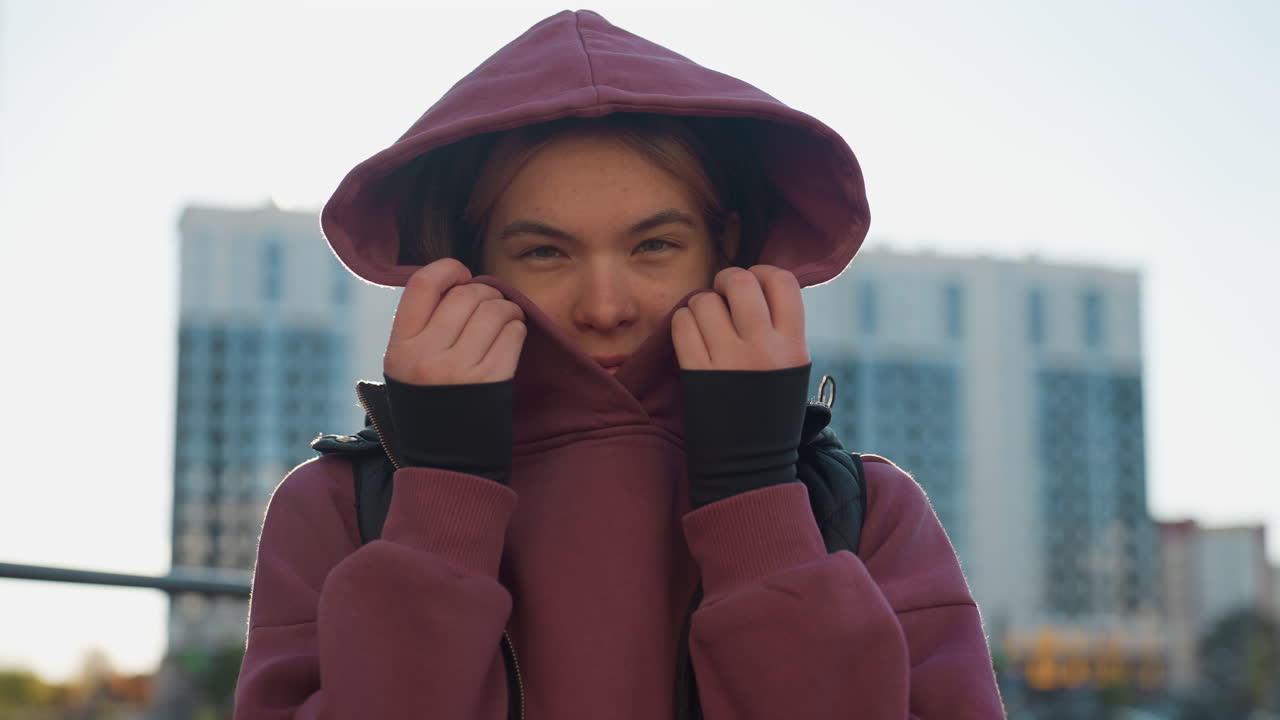 Jogger covers face with maroon hoodie and black vest under bright sun in urban park, playful shy glance from hooded athlete pausing between training sets by pull up bars in city outdoor workout