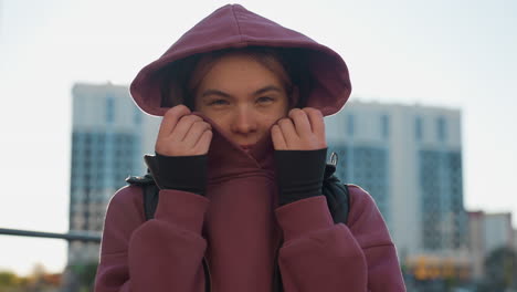 jogger covers face with maroon hoodie and black vest under bright sun in urban park, playful shy glance from hooded athlete pausing between training sets by pull up bars in city outdoor workout