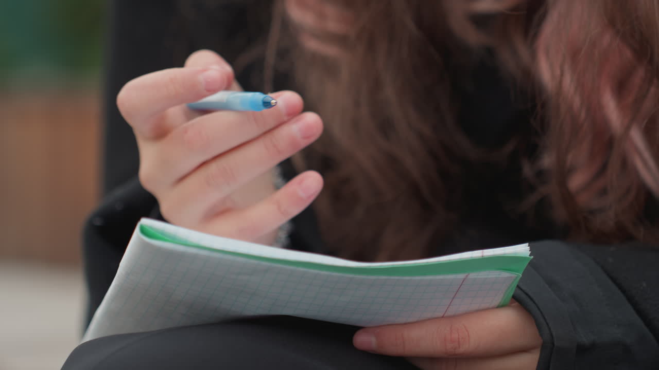 student writing notes with blue pen on spiral notebook outdoors, long hair, cozy coat, pearl bracelet, closeup hands making thoughtful gestures, focused study vibe under soft natural light