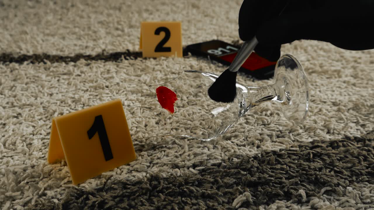 Close up view of a police forensic investigator dusting fingerprints from a borken wine or beer glass on carpet with yellow crime scene markers next to it. Phone in the blurry background calling 911