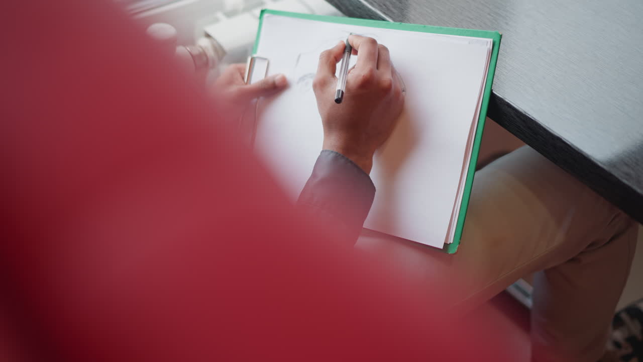 Immigrant sketching vehicle with black pen on white paper attached to green folder, viewed from above near window, suggesting creativity, technical drawing, concept development