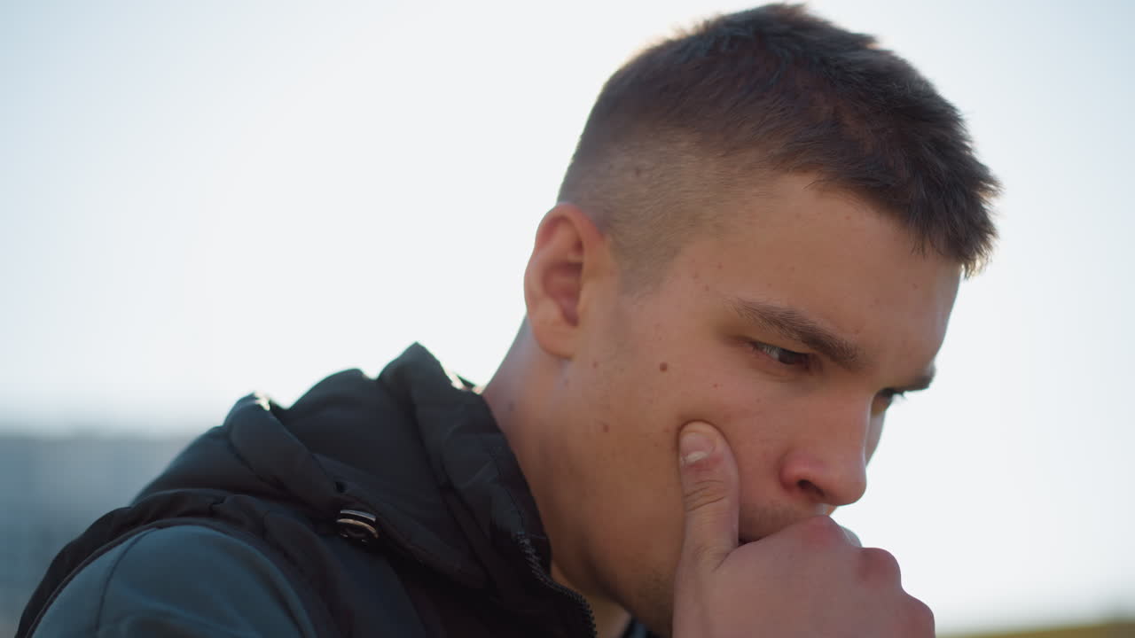 close up side view of white boy holding vape device near lips while inhaling during sunny day wearing black vest and t-shirt focused expression with blurred background and bright natural lighting
