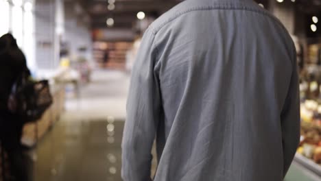 Traking-rare-footage-of-a-man-in-blue-shirt-pushing-the-shopping-trolley-the-supermarket-entry.-Blurred-background-of-the-food-shelves-in-grocery-department