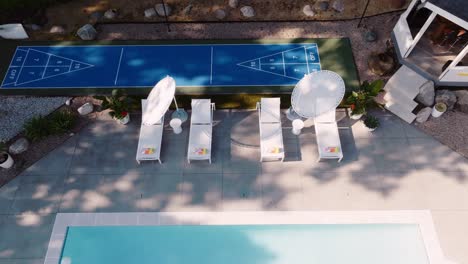 a panning aerial drone shot of a backyard pool seating area with white lounge chairs and a shuffleboard court in the background