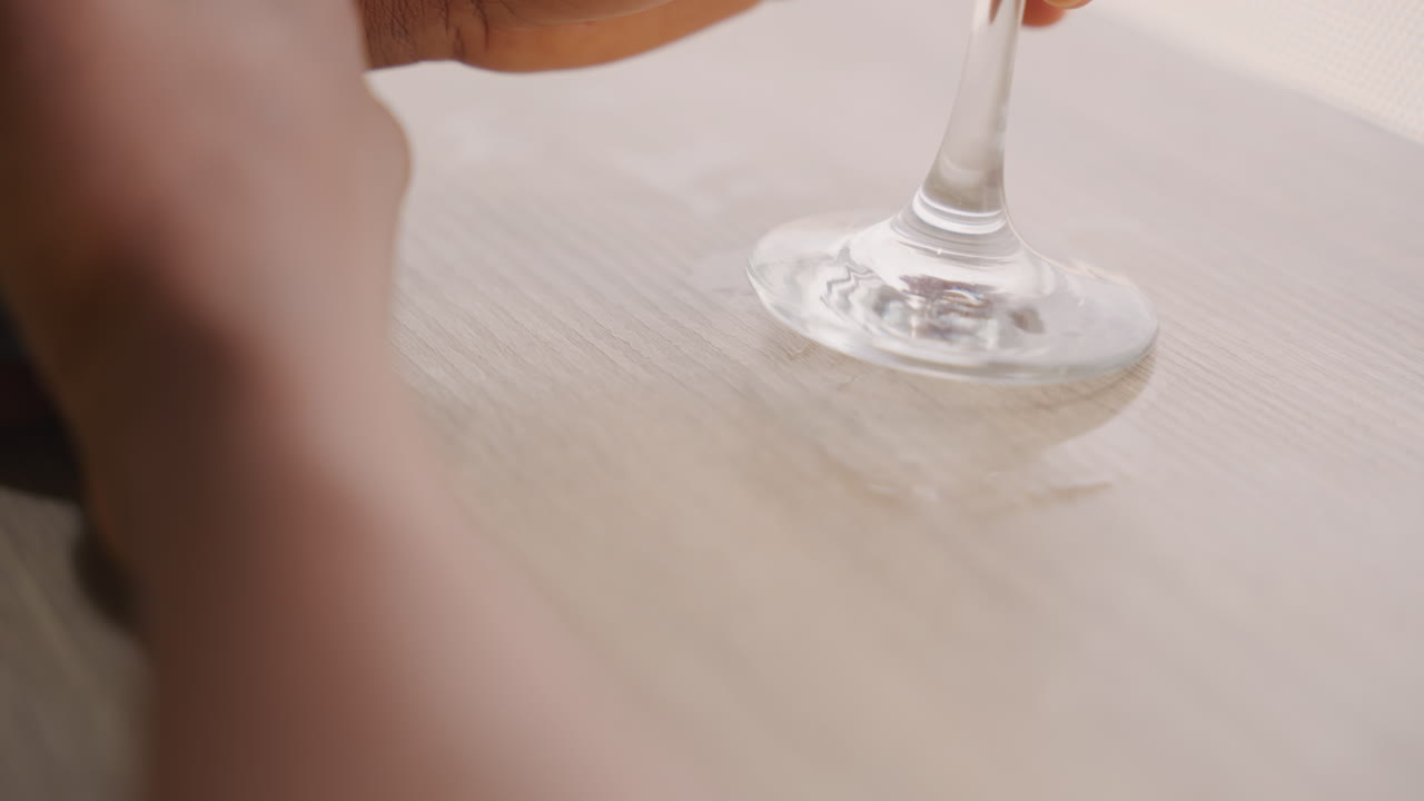 Delicato calice da vino su tavolo di legno, stelo appoggiato con un leggero anello di condensa, mano che tiene fermo il bicchiere, piccola fuoriuscita di liquido e increspatura, luce diurna soffusa e atmosfera intima da appuntamento serale