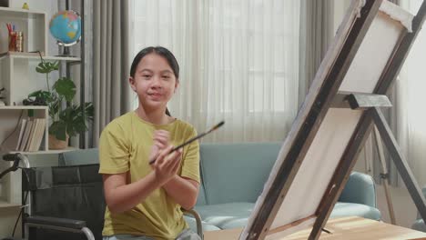 asian artist girl in wheelchair holding paintbrush and cross his arms and smile while painting on the canvas