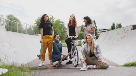 Retrato-De-Amigas-Con-Patinetas-Y-Bicicletas-Paradas-En-Un-Parque-De-Patinaje-Urbano