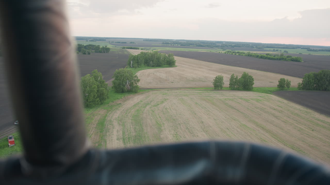 Overhead view of expansive open farmland patchwork from hot air balloon basket edge, fields in various growth stages, tree clusters dotted along horizon