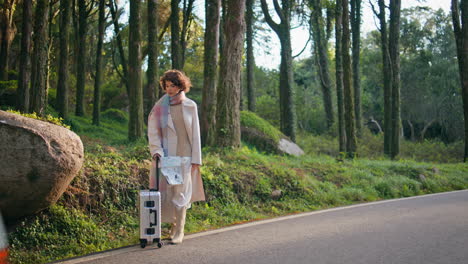 traveler standing roadside suitcase at autumnal forest. woman waiting journey