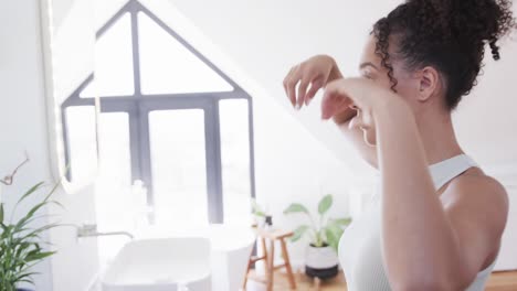 Glückliches-Gemischtrassiges-Lesbisches-Paar,-Das-Seine-Haare-Und-Sein-Gesicht-Im-Spiegel-Im-Sonnigen-Badezimmer-überprüft,-Zeitlupe