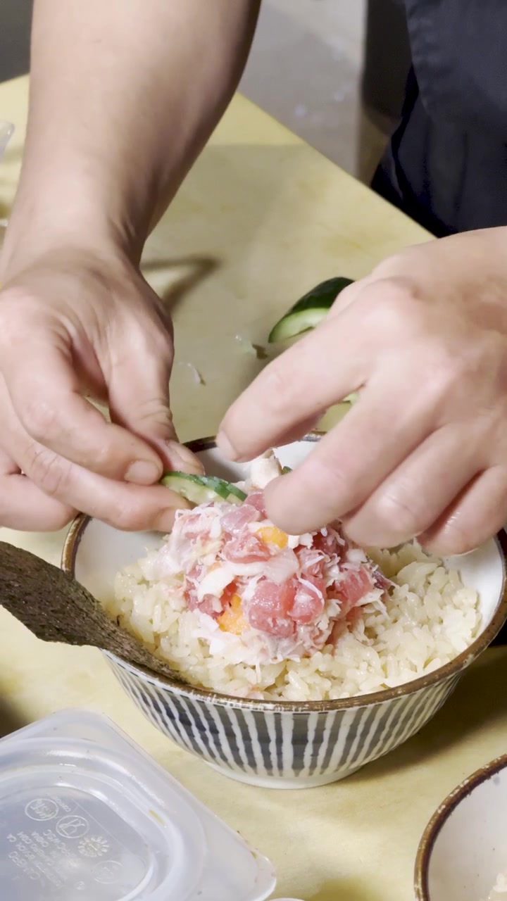 Close-up tangan koki menghias mangkuk nasi salmon tuna di bawah pencahayaan dapur atas yang terang.