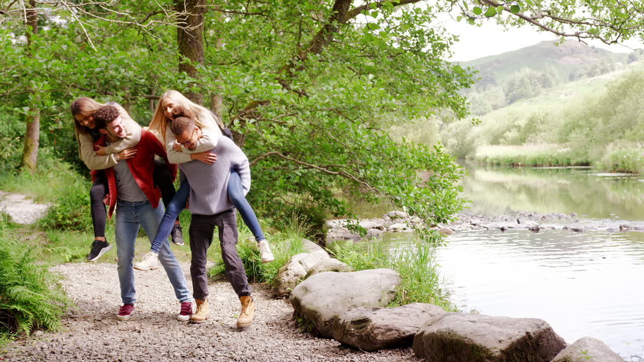 Four young adult friends during a hike, men piggybacking their girlfriends, handheld
