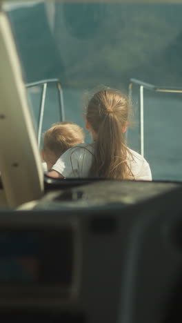 girl with waving hair hugs little brother sitting on sailboat deck view from cockpit. lovely children look in distance sailing yacht on summer vacation