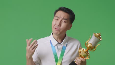 cerca de preguntarse hombre asiático con una medalla de oro mirando el trofeo dudosamente en el fondo de la pantalla verde en el estudio