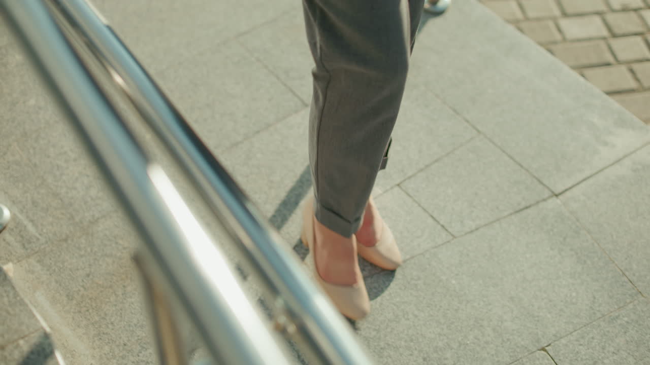 Top down view of lady in heels and blazer walking along tiled pavement bordered with iron railing in bright daylight, showcasing confident stride and urban style in professional outdoor environment