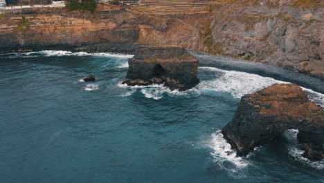 Los-Roques-Beach,-Tenerife:-aerial-view-travelling-out-of-the-two-rock-formations-of-the-famous-Tenerife-beach