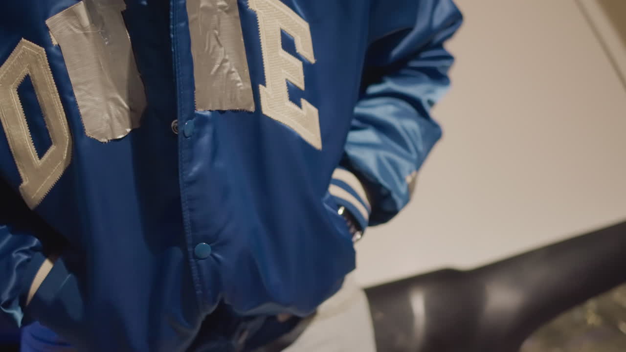 Conducción nocturna: primer plano de un conductor ajustándose una chaqueta azul de satén, con las manos subiendo la cremallera y metiendo la chaqueta dentro, junto a un coche aparcado bajo farolas de neón; reloj de pulsera visible, ambiente informal de ropa urbana, bokeh cinematográfico y ambiental