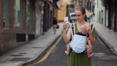 while carrying her infant in a kangaroo backpack, a young woman documents her travel experiences using her mobile phone. as she walks, she intermittently checks the screen