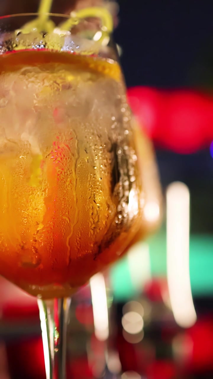 Un barista aggiunge una scorza di agrumi a un cocktail freddo e arancione in un bicchiere con stelo. Luci del bar vivaci e colorate creano un'atmosfera notturna festosa. Profondità di campo ridotta.