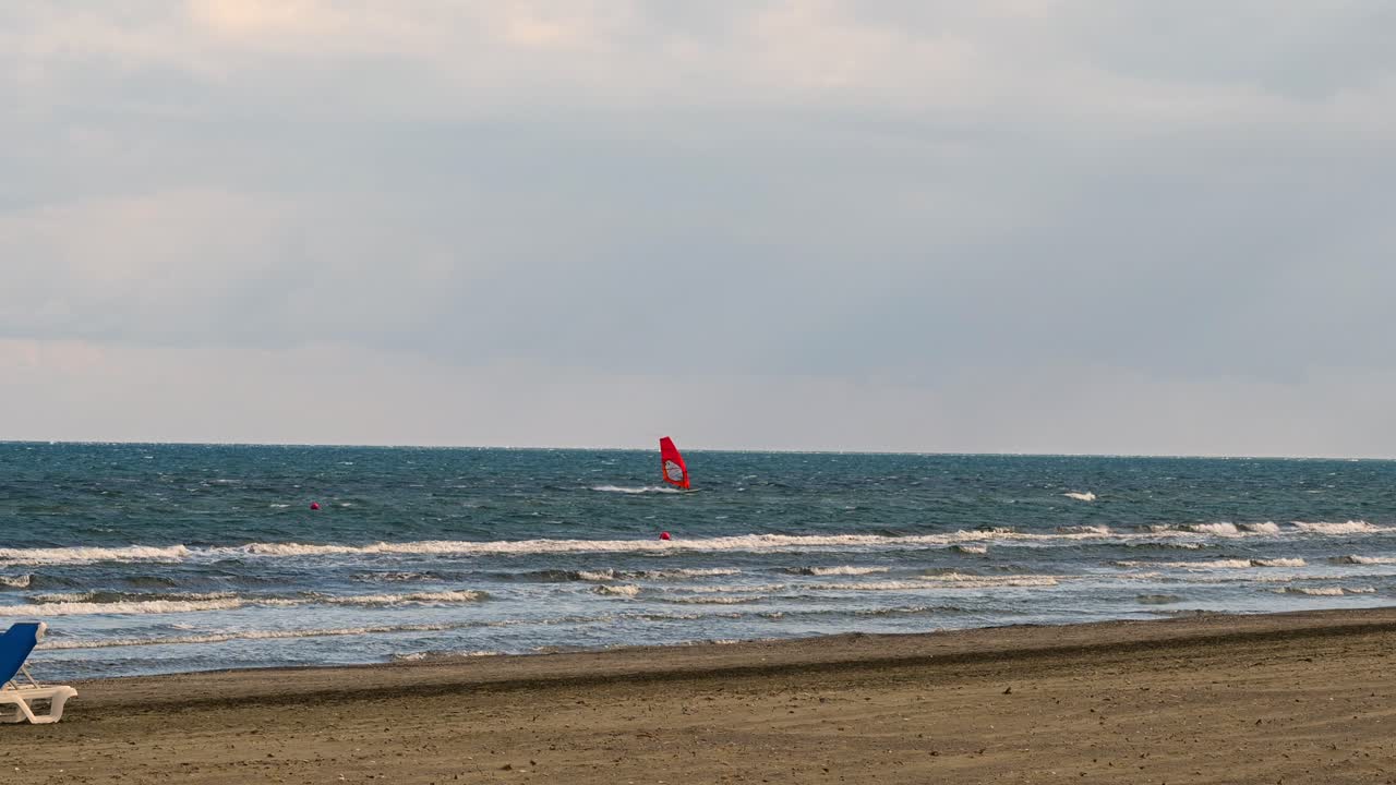 Distant view of a windsurfer with a bright red sail gliding across the Mediterranean Sea off the coast of Larnaca, Cyprus, with waves breaking on the sandy beach