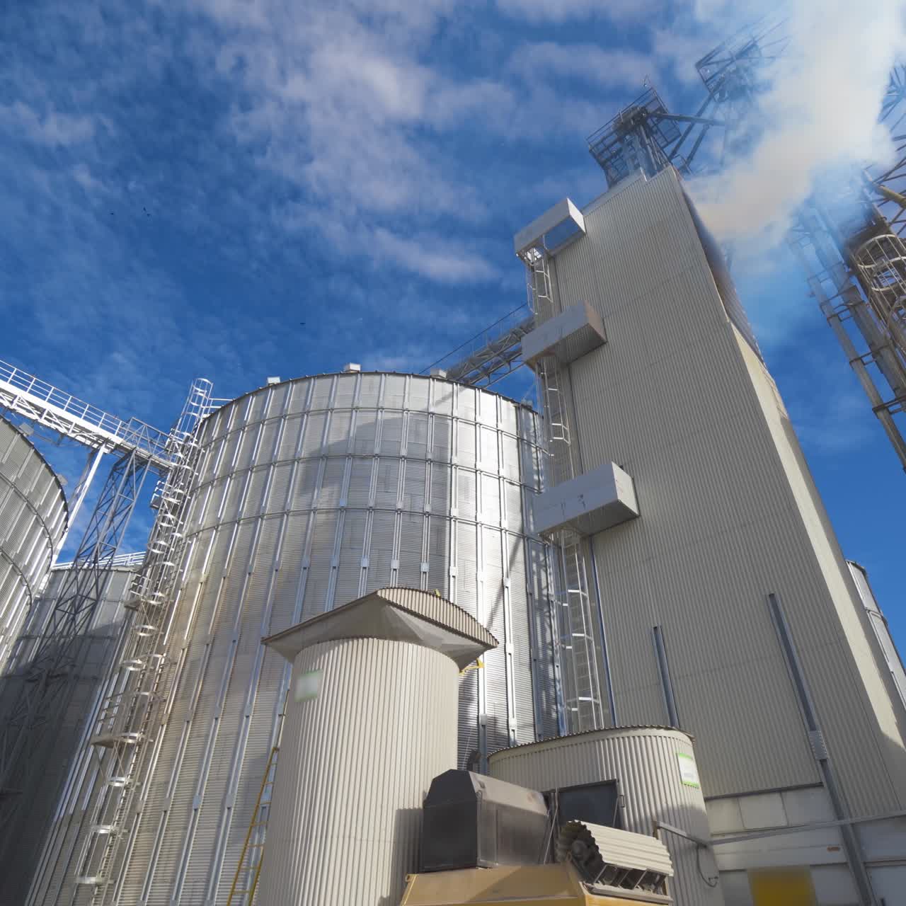 Modern grain elevators outdoors. Agricultural complex for grain processing. White smoke from the industrial plant on blue sky background